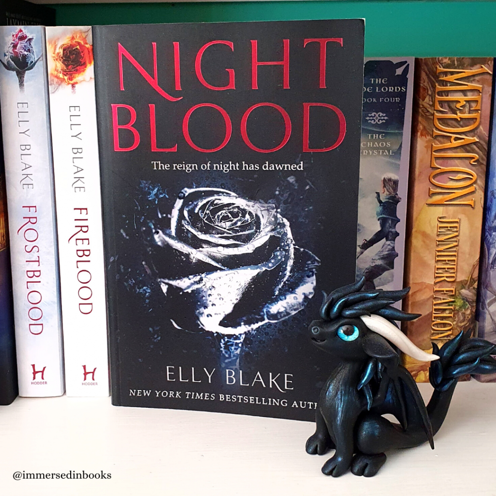 Nightblood by Elly Blake is situated in the middle of the photo, on a white bookshelf and in front of Blake's other books to the left, with Jennifer Fallon's books to the right. Just in front and to the right of Nightblood is a black dragon with blue touches, by Dirty Paws Australia.