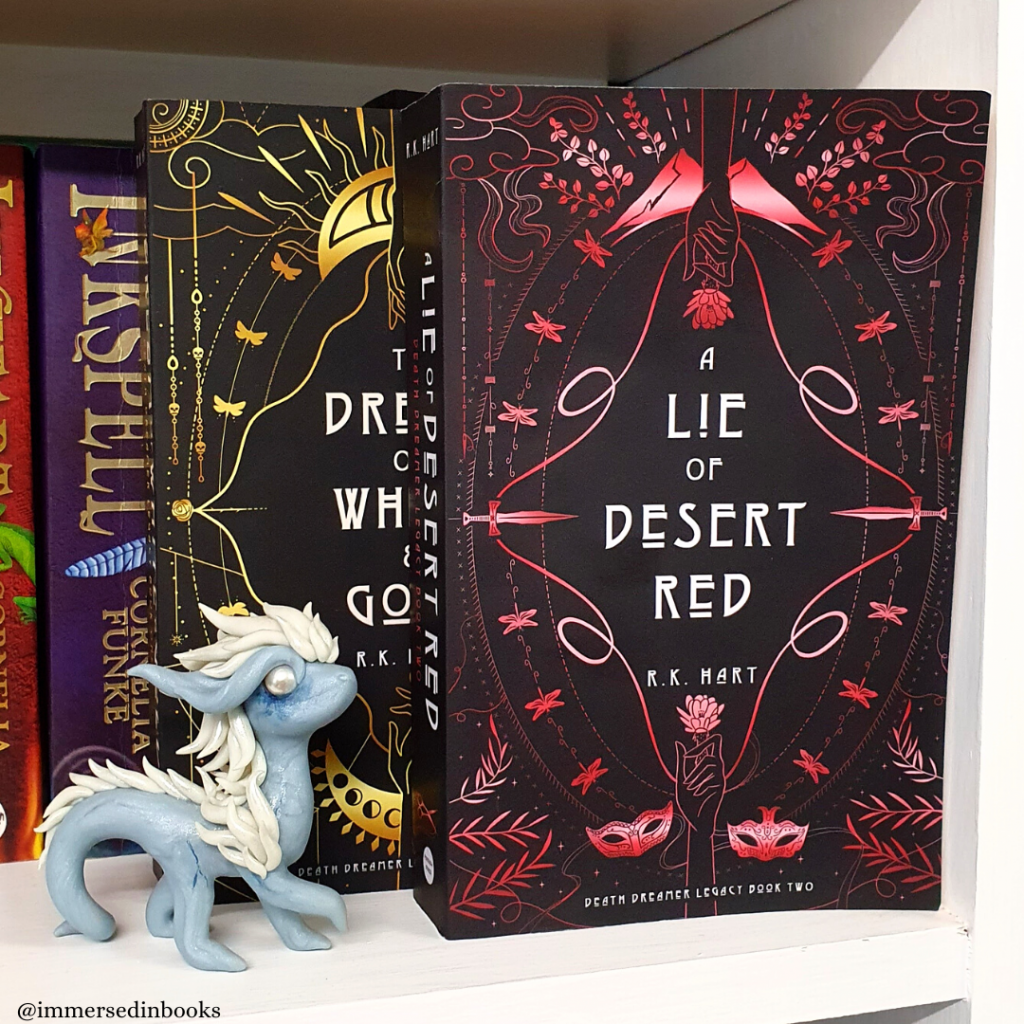 Paperback of A Lie of Desert Red by R. K. Hart is to the right of the image, on a white bookshelf and in front of the first book of the series (To Dream of White & Gold), as well as Cornelia Funke's Inkworld trilogy. The ghostly, glow in the dark blue and white dragon in front of the books is by Dirty Paws Australia.