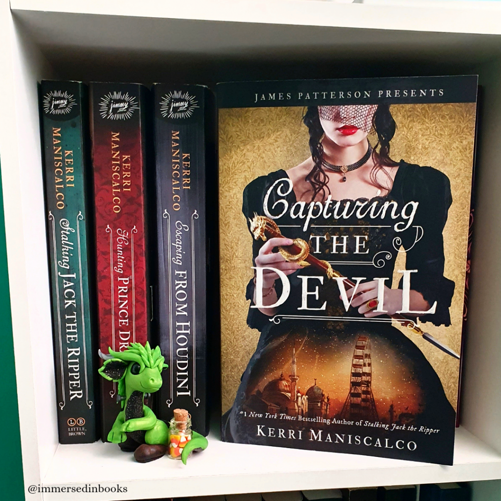 Paperback copy of Capturing the Devil by Kerri Maniscalco is to the right of the 3 other books in this series on a white bookshelf. A Frankenstein monster green dragon by Dirty Paws Australia sits to the left of the book, with mini candy corn in a tiny jar.