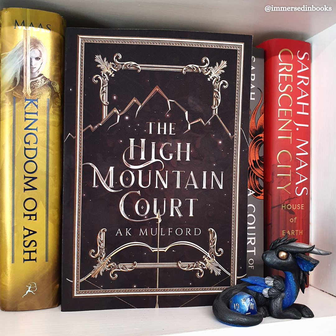 A paperback copy of The High Mountain Court by A. K. Mulford is sitting on a white bookshelf, in front of Sarah J. Maas' books. A blue and black night dragon with a golden eye and blue dice by Dirty Paws Australia is sitting to the right of the book.