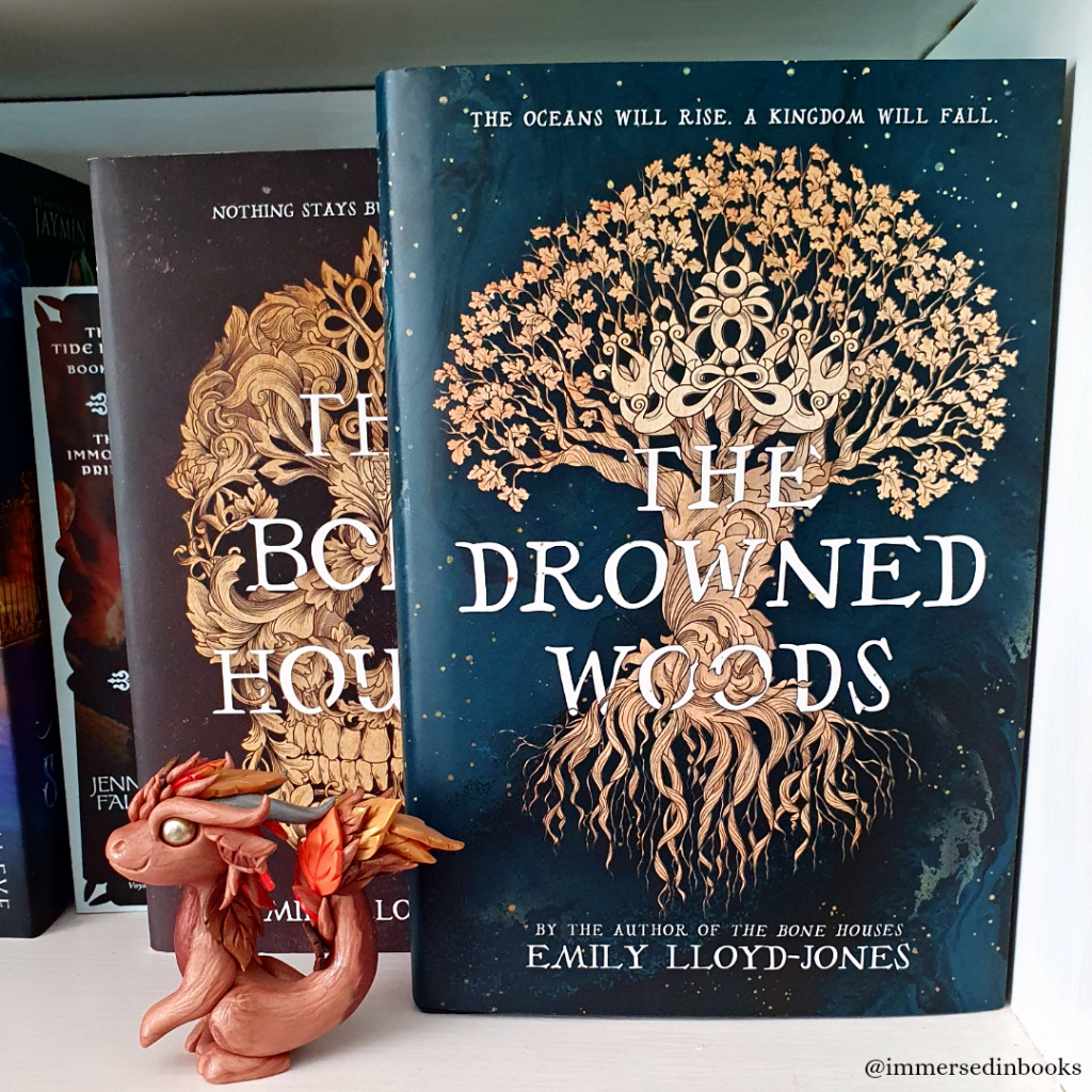 A hardback copy of The Drowned Woods by Emily Lloyd-Jones is just in front and to the right of one of her other books, The Bone Houses. They sit on a white bookshelf and a autumnal dragon by Dirty Paws Australia sits to the left of The Drowned Woods and in front of The Bone Houses.