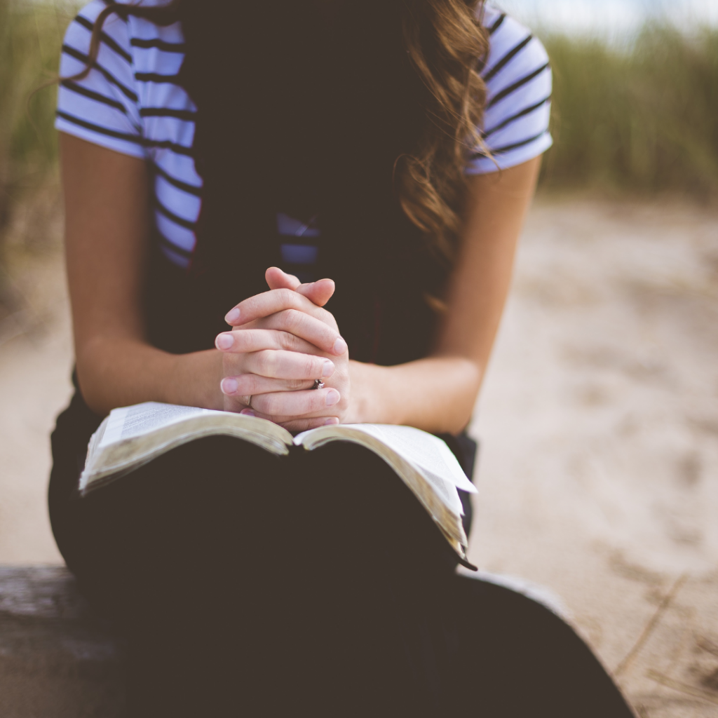 November Book Goals includes lots of reading! The image is of a dark brown, long haired Caucasian female sitting on a beach, with a book on her lap. Her hands are folded over the open pages.