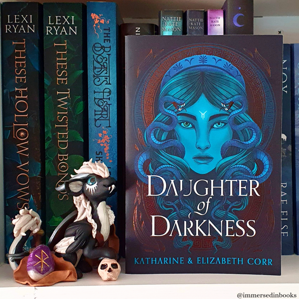 A paperback copy of Daughter of Darkness by Katharine and Elizabeth Corr is on a white bookshelf, in front of books by Nattie Kate Mason, Rae Else and Lexi Ryan. A Freya inspired polymer clay dragon by Dirty Paws Australia is to the left of the book.