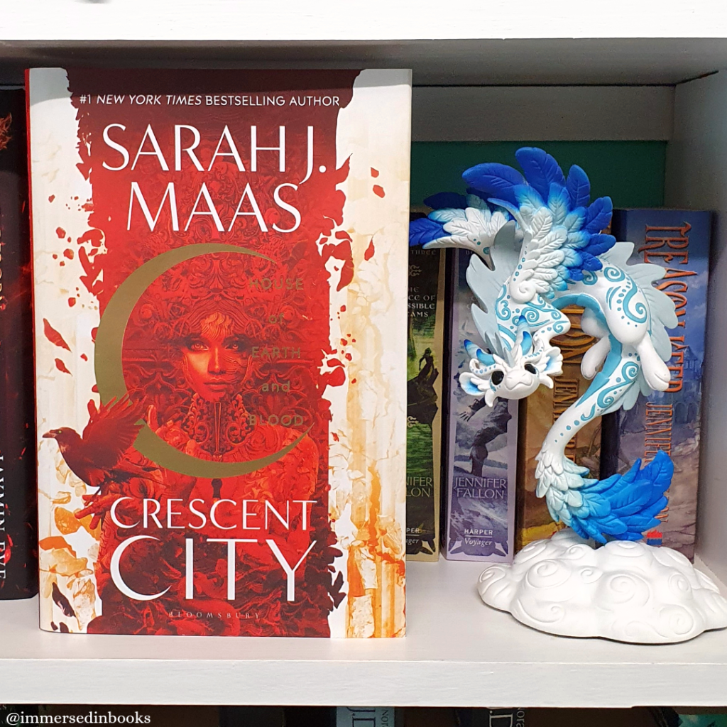 Hardcover of House of Earth and Blood by SJM is on a white bookshelf, to the left of the image and in front of books by Jennifer Fallon. To the right of the book is the Swift dragon by Dragons & Beasties.